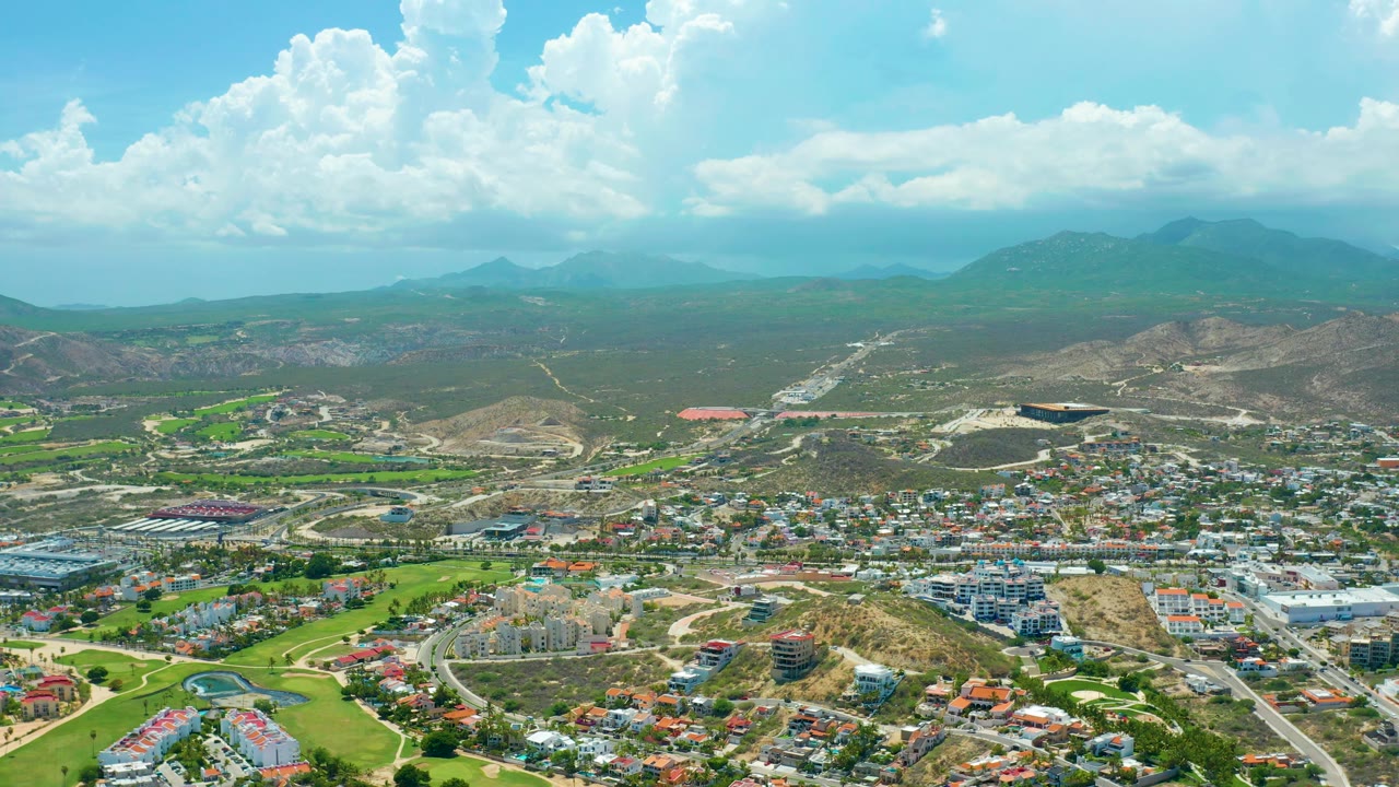 Mid-clip aerial frame of San Jose del Cabo showing roads crossing the town, green fields at left, brown hills beyond, cloud shadows, and no visible people.