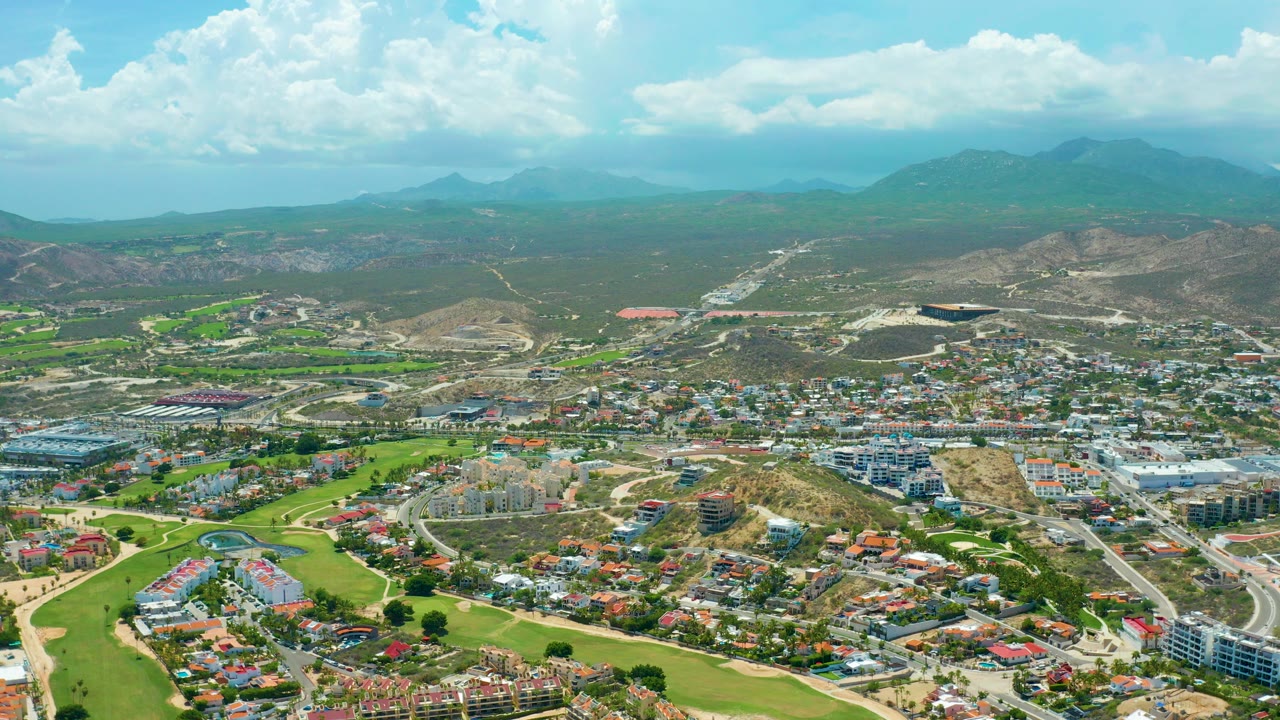 Wide aerial frame over San Jose del Cabo showing green golf fairways, residential blocks, pale roads, dry hills, distant mountains, and no visible people.