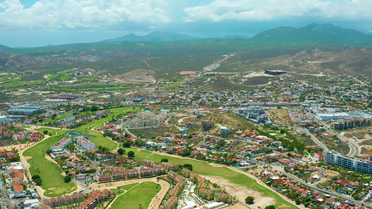 Opening aerial frame over San Jose del Cabo showing green fairways, residential blocks, dry hills, distant mountains, broad cloud cover, and no visible people.
