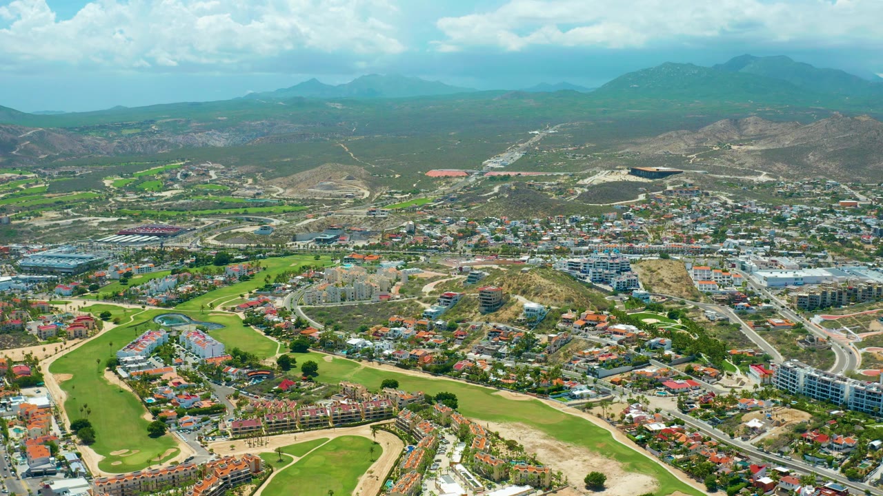 Two-second frame from San Jose del Cabo showing a golf-course greenbelt, hillside residential development, pale roads, distant ridges, bright clouds, and no visible people.