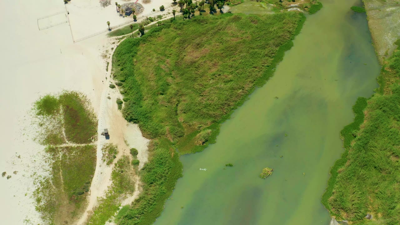 Aerial frame in San Jose Del Cabo with greenish water running between sandy shore and thick vegetation, while the small white boat sits slightly farther along the channel and no visible people appear.