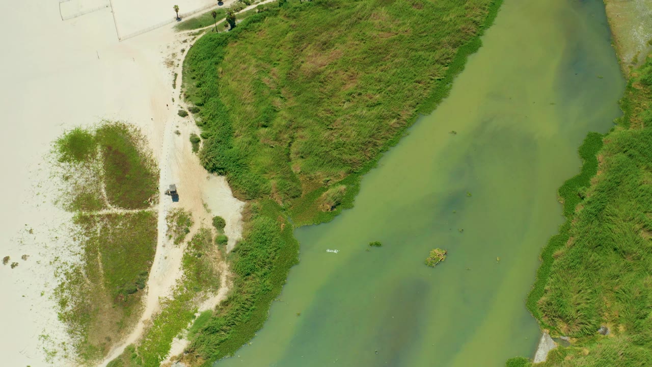 Top-down San Jose Del Cabo river view with the green channel widening through the frame, sand and footpaths at left, dense grass at right, and a small white boat visible with no visible people.