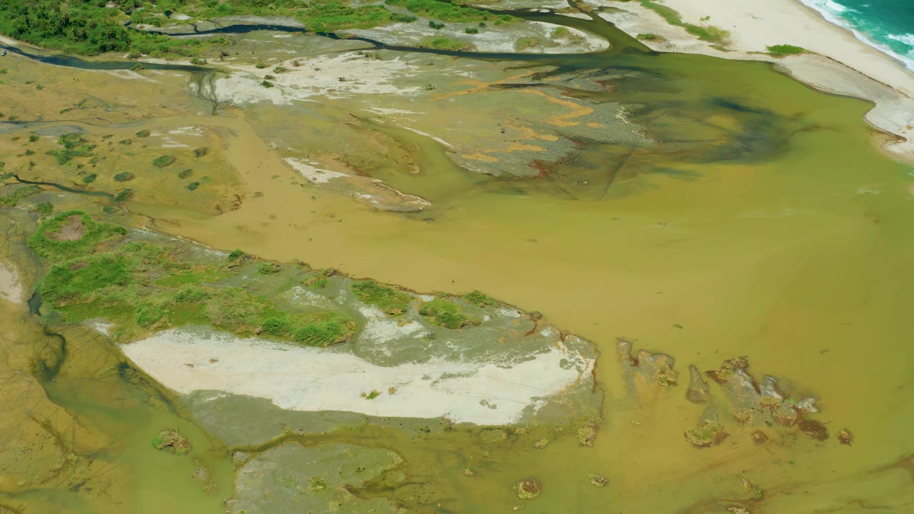 Second aerial frame in San Jose del Cabo showing the sandy lower edge, broad green-brown water, darker wetland streaks, scattered vegetation, and no visible people.