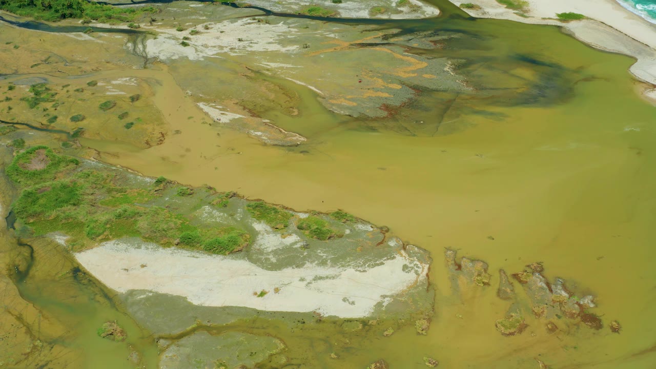 Mid-clip aerial frame in San Jose del Cabo with a large green water channel, pale sandy flats across the bottom, darker wet areas near the top, and no visible people.