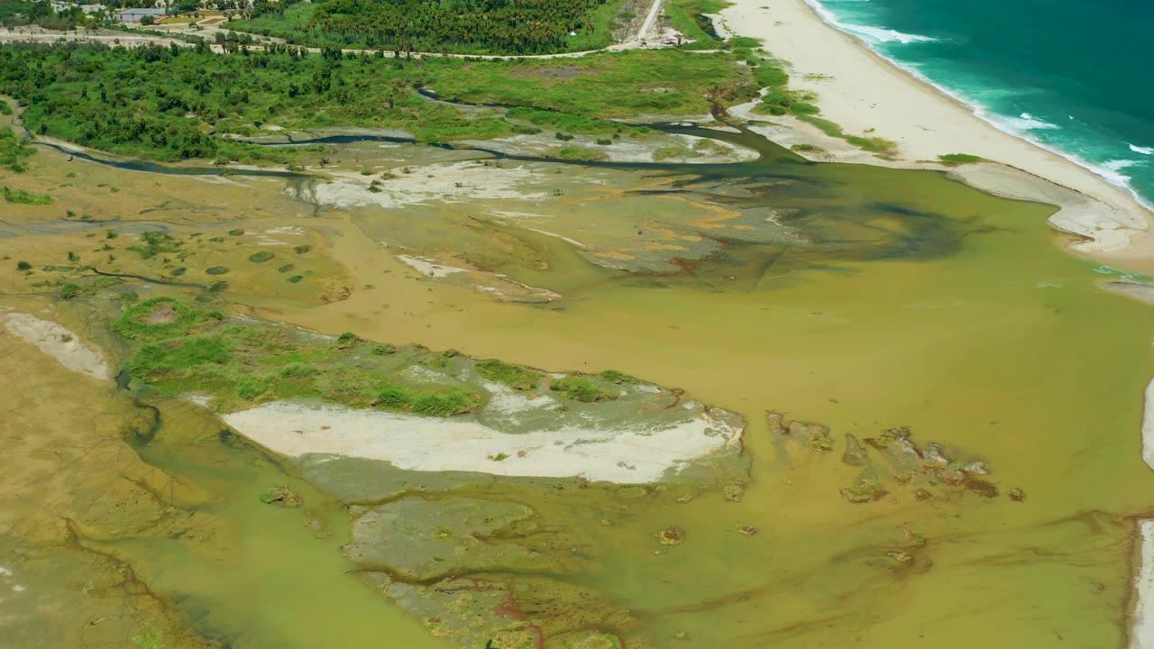 Aerial view in San Jose Del Cabo with brown estuary channels crossing green wetland patches toward pale sand and ocean surf, with no visible people.