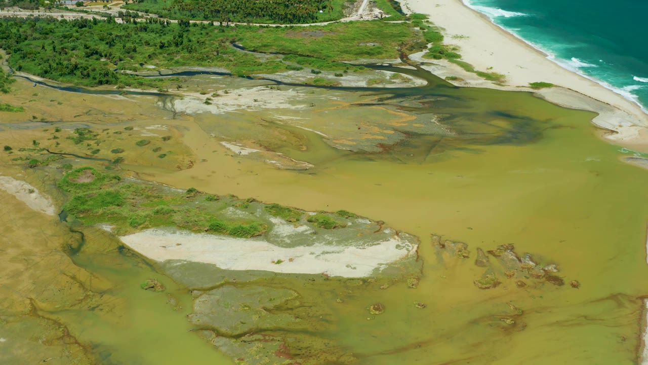 San Jose Del Cabo aerial frame showing a muddy lagoon area with sand flats below a bright beach and blue-green ocean waves, with no visible people.