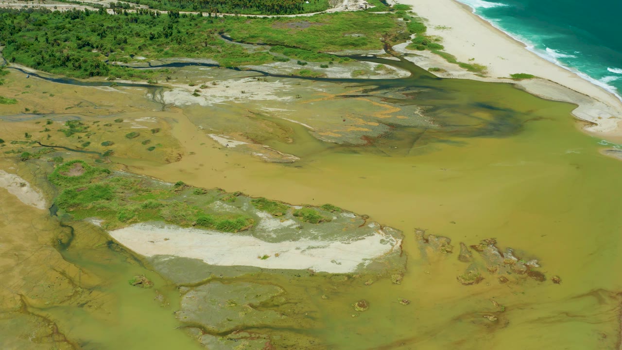 Overhead San Jose Del Cabo view with yellow-brown estuary water surrounding sand islands and green shoreline vegetation near ocean waves, with no visible people.