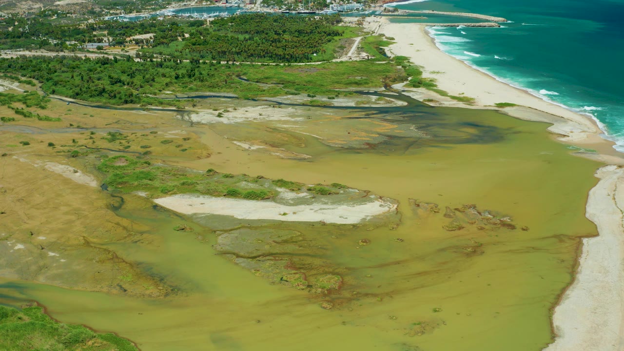 Aerial frame over San Jose del Cabo wetlands with green-brown water, sandy beach, turquoise surf, inland vegetation, roads, and a tiny beach figure near the early shoreline view.