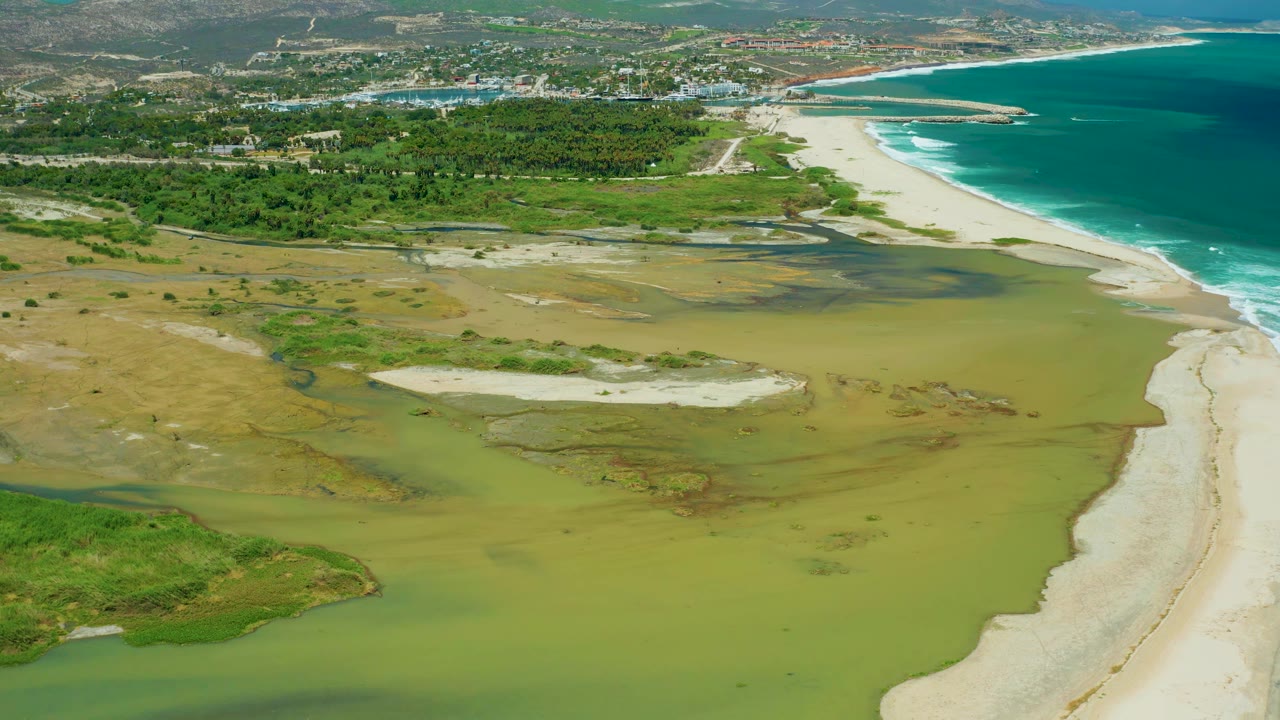 Aerial view in San Jose Del Cabo with shallow green wetland water across the lower frame, a white sand strip on the right, turquoise ocean at the top right, and no visible people.