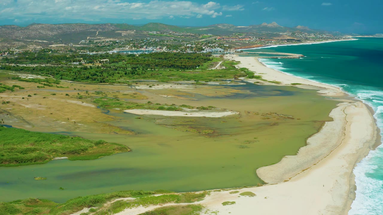 San Jose del Cabo aerial view with the lagoon spread across the lower frame, white sand curving along the right edge, surf lines on the ocean, coastal development in the distance, and no visible people.