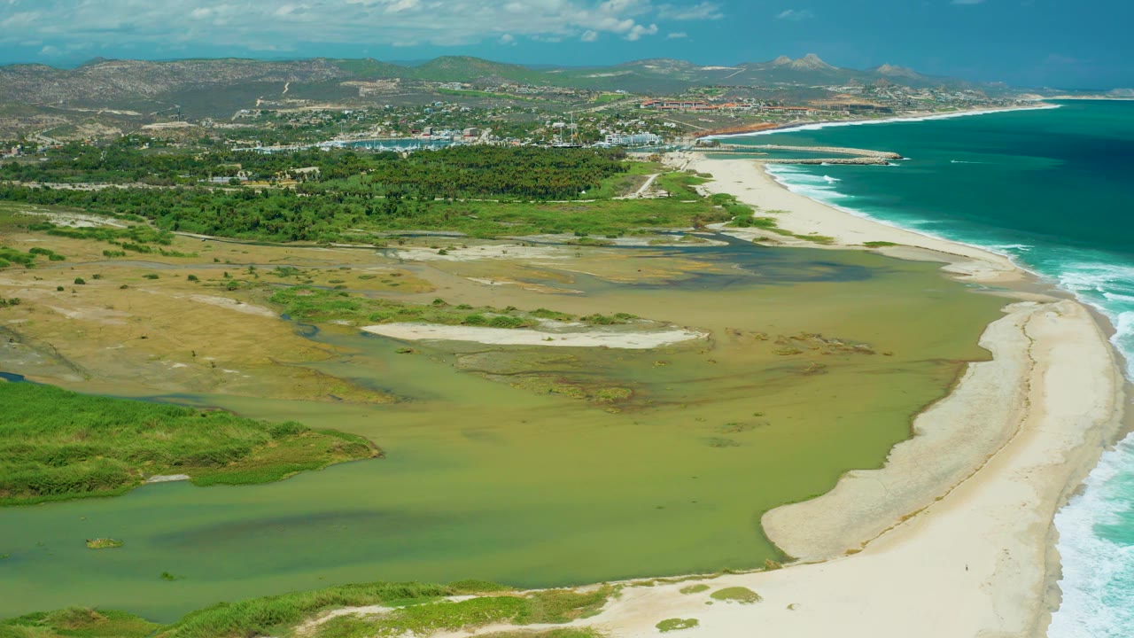 Aerial frame over San Jose del Cabo where green wetland water fills the lower half, a bright sand spit divides lagoon from beach, the ocean runs along the right side, and no visible people appear.