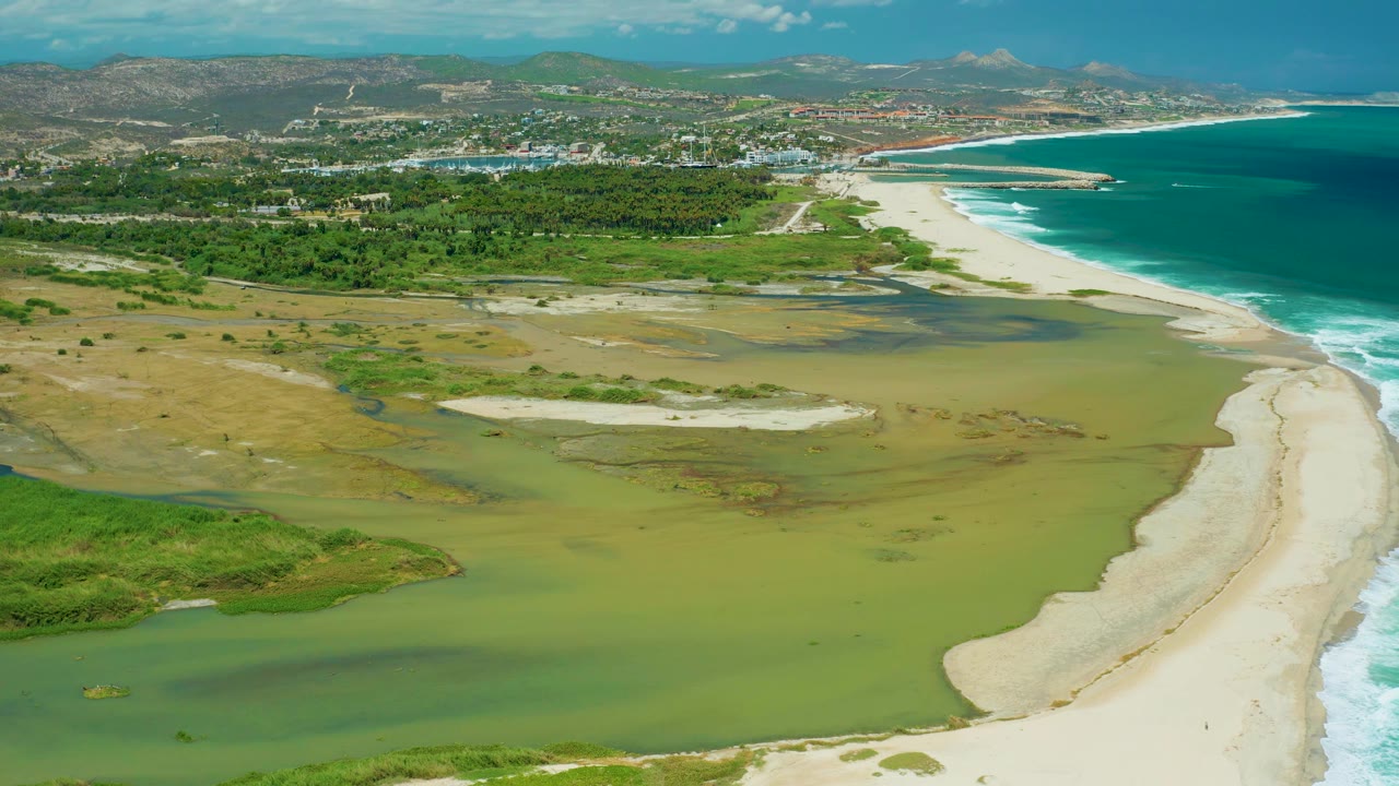San Jose del Cabo aerial view with darker cloud shadow across parts of the lagoon, tan wetland flats at left, bright beach and surf at right, distant buildings and mountains, and no visible people.