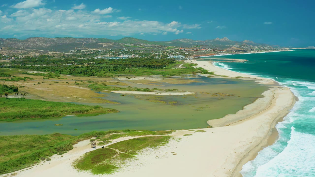 Aerial frame over San Jose del Cabo showing the sandbar more centered between the green estuary lagoon and white ocean break, with hills and town structures in the background and no visible people.