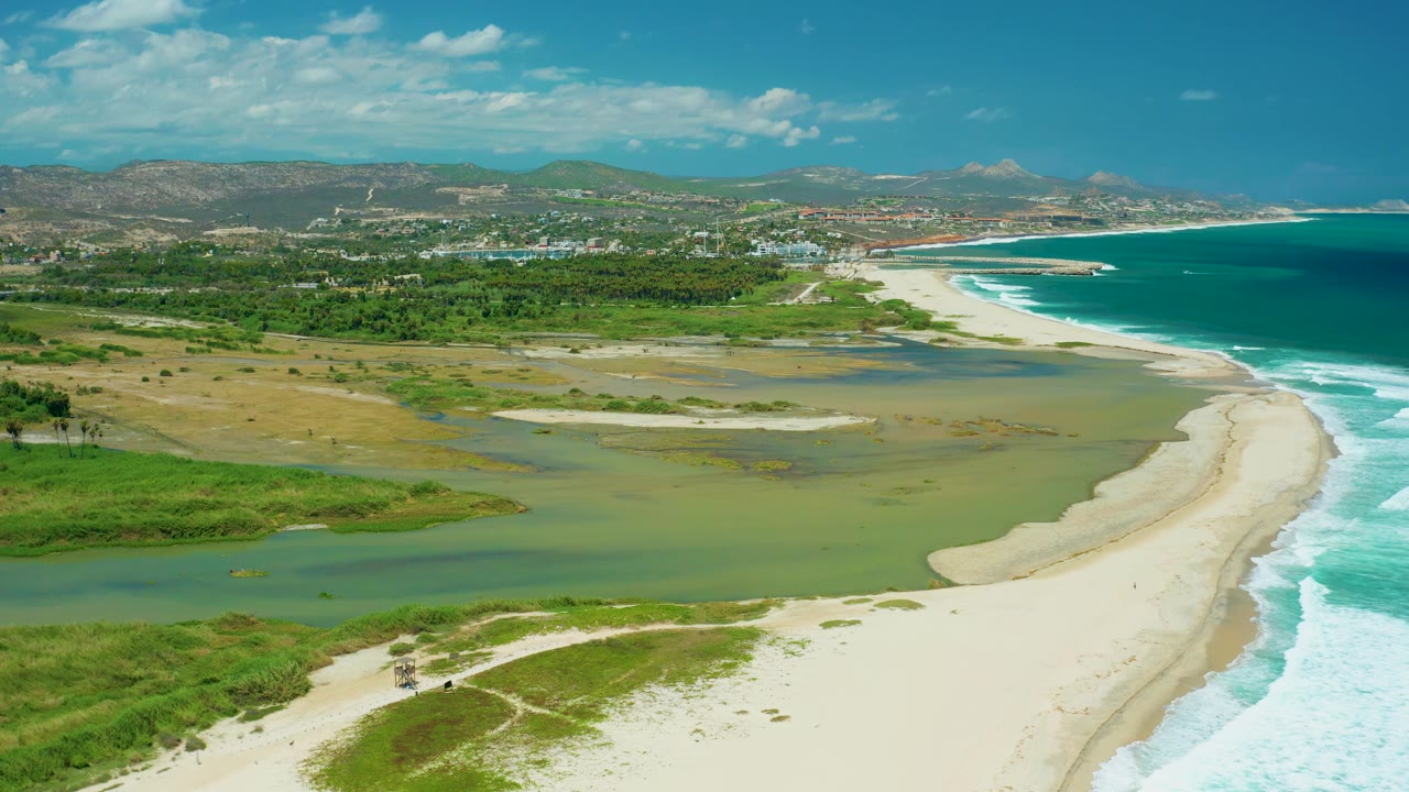 Wide drone view of the San Jose del Cabo coastline, with shallow green estuary water in the center, white beach and surf on the right, low vegetation below, and no visible people.