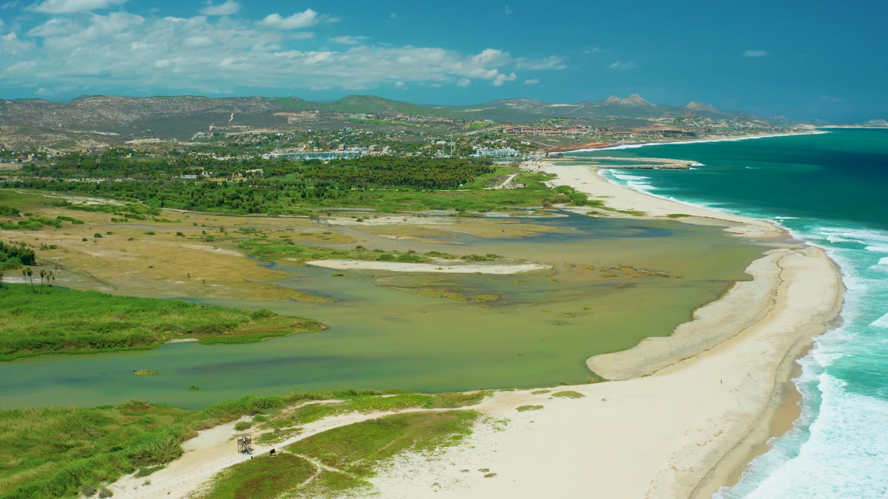 San Jose del Cabo aerial view with pale estuary flats spread across the midground, a bright beach along the right side, breaking ocean waves, green foreground plants, and no visible people.