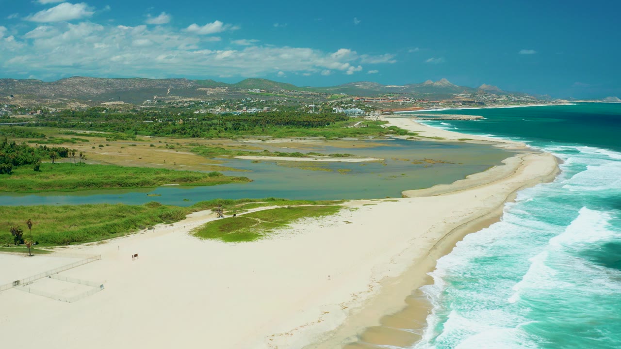 Aerial view over San Jose del Cabo showing a pale sandy beach beside a shallow estuary, turquoise surf along the right side, distant town and hills, and no visible people.