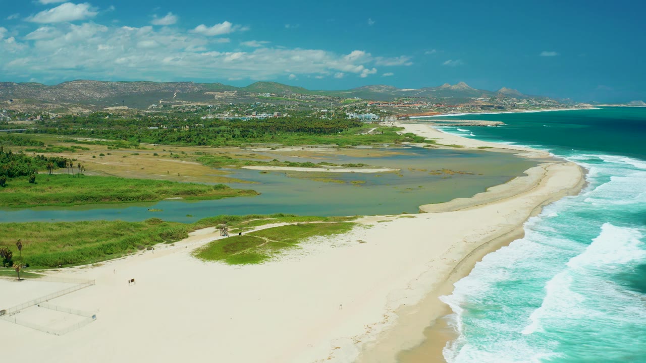 Aerial view in San Jose del Cabo with the estuary stretching across the midground, bright beach sand in the foreground, ocean surf on the right, green banks and hills beyond, and no visible people.