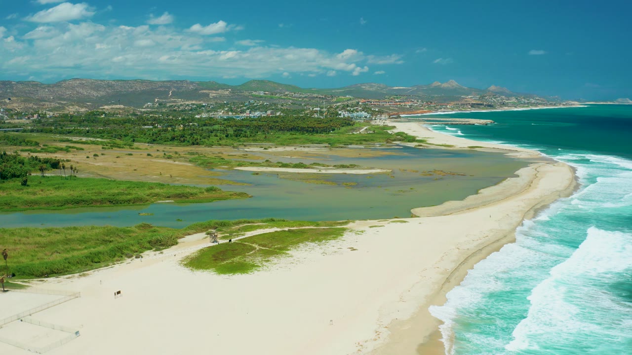 Aerial shoreline view with shallow estuary, beach path, ocean waves, and hills.