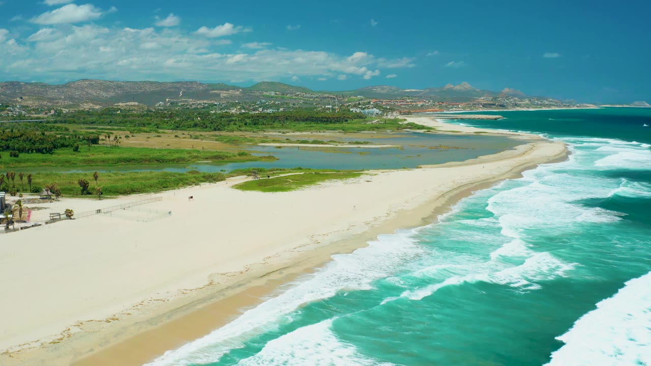Aerial view in San Jose del Cabo with white sand curving beside foamy turquoise waves, lagoon water set inland, green shore vegetation, and no visible people.