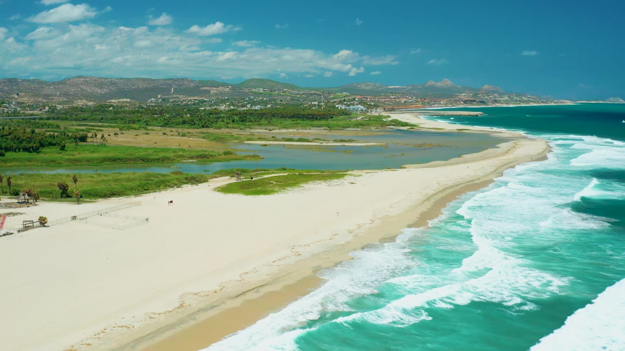 A high aerial frame over San Jose del Cabo shows a broad empty-looking beach, turquoise waves, lagoon channels, green vegetation, distant town structures, hills, and no visible people.
