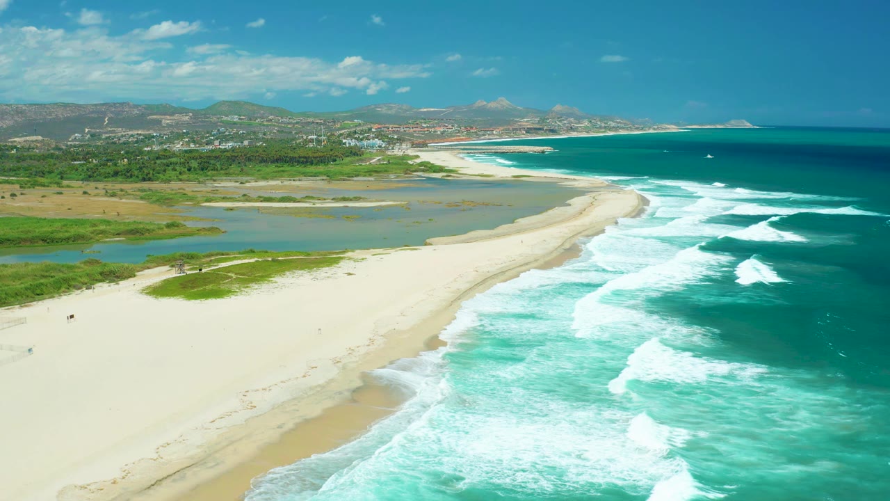 Wide aerial frame in San Jose del Cabo with the lagoon more centered, the sandbar curving toward the horizon, ocean waves on the right, and no visible people.