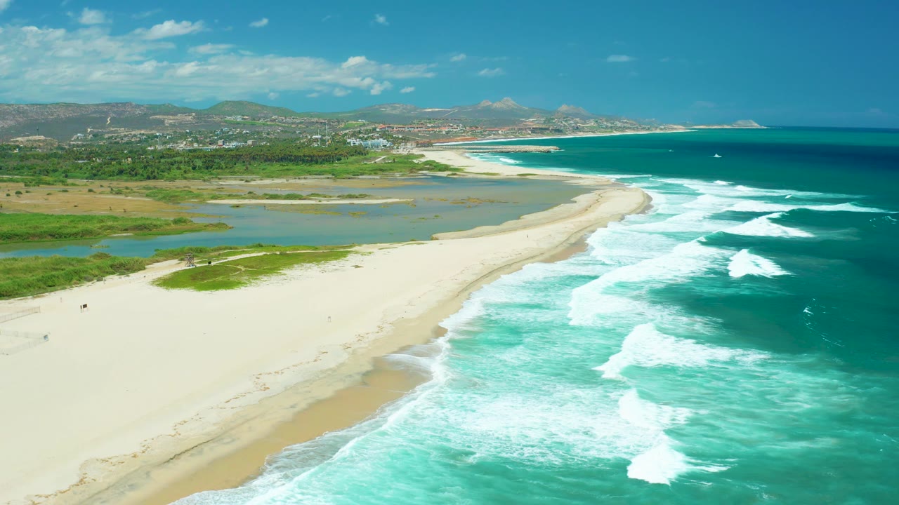 Aerial frame over San Jose del Cabo with bright surf pushing toward the beach, lagoon water behind the sand, distant buildings, mountains, and no visible people.