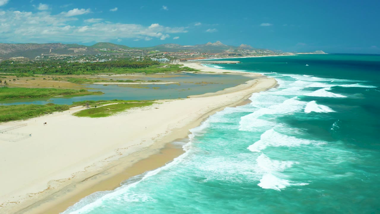 San Jose del Cabo aerial frame showing the beach curve from lower left to center, lagoon and greenery to the left, ocean surf to the right, and no visible people.