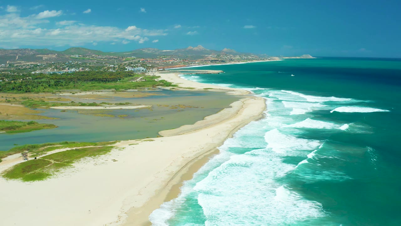 Aerial San Jose Del Cabo beach view with a long white surf line, calm lagoon water behind the sand, green shoreline patches, distant buildings, and no visible people.