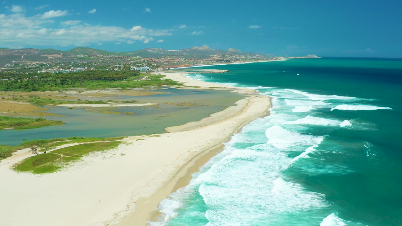 San Jose Del Cabo aerial coastline frame with brighter surf foam spread across the lower right, lagoon water to the left of the sandbar, distant hills and buildings, and no visible people.