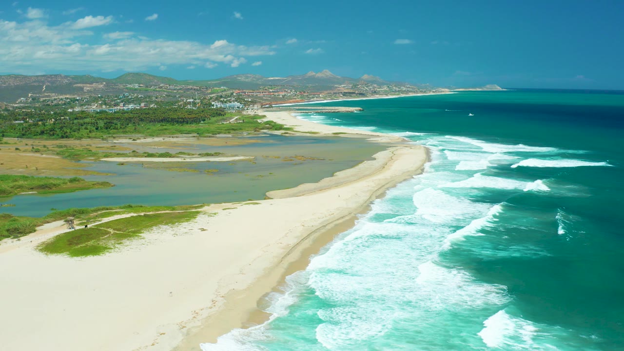 High view over San Jose Del Cabo shoreline with white waves breaking close to the beach, lagoon and marshy green areas behind the sand, distant town and hills, and no visible people.