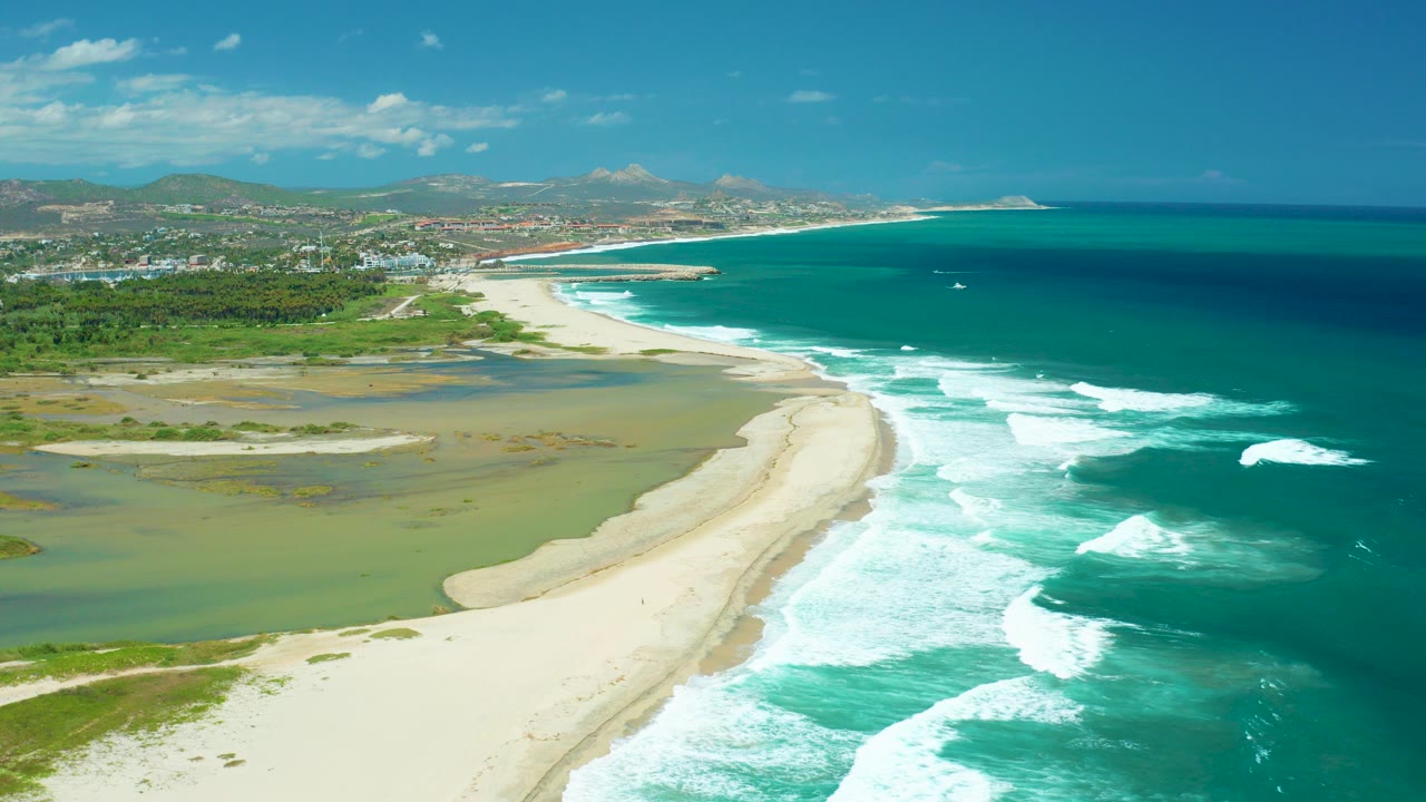 Aerial view over San Jose Del Cabo coastline with a curved sandy beach, turquoise waves, shallow inland water, distant buildings, mountains, scattered clouds, and no visible people.