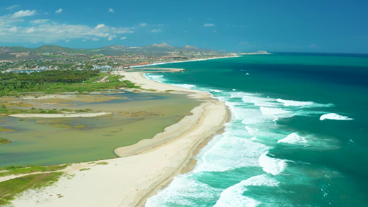 Wide drone view of beach, waves, inland water, greenery, distant town, and mountains.