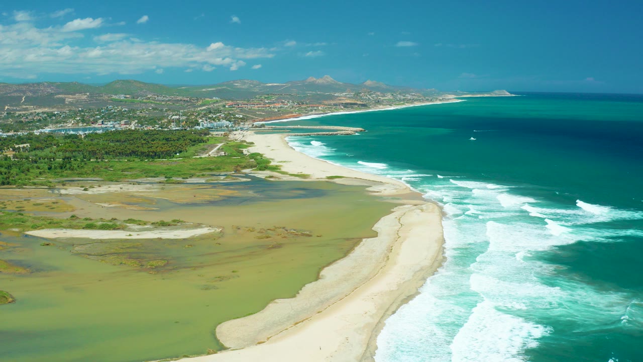 Aerial opening frame over San Jose del Cabo coastline showing a curved sand beach between green lagoon water and turquoise ocean surf, with distant hills and no visible people.
