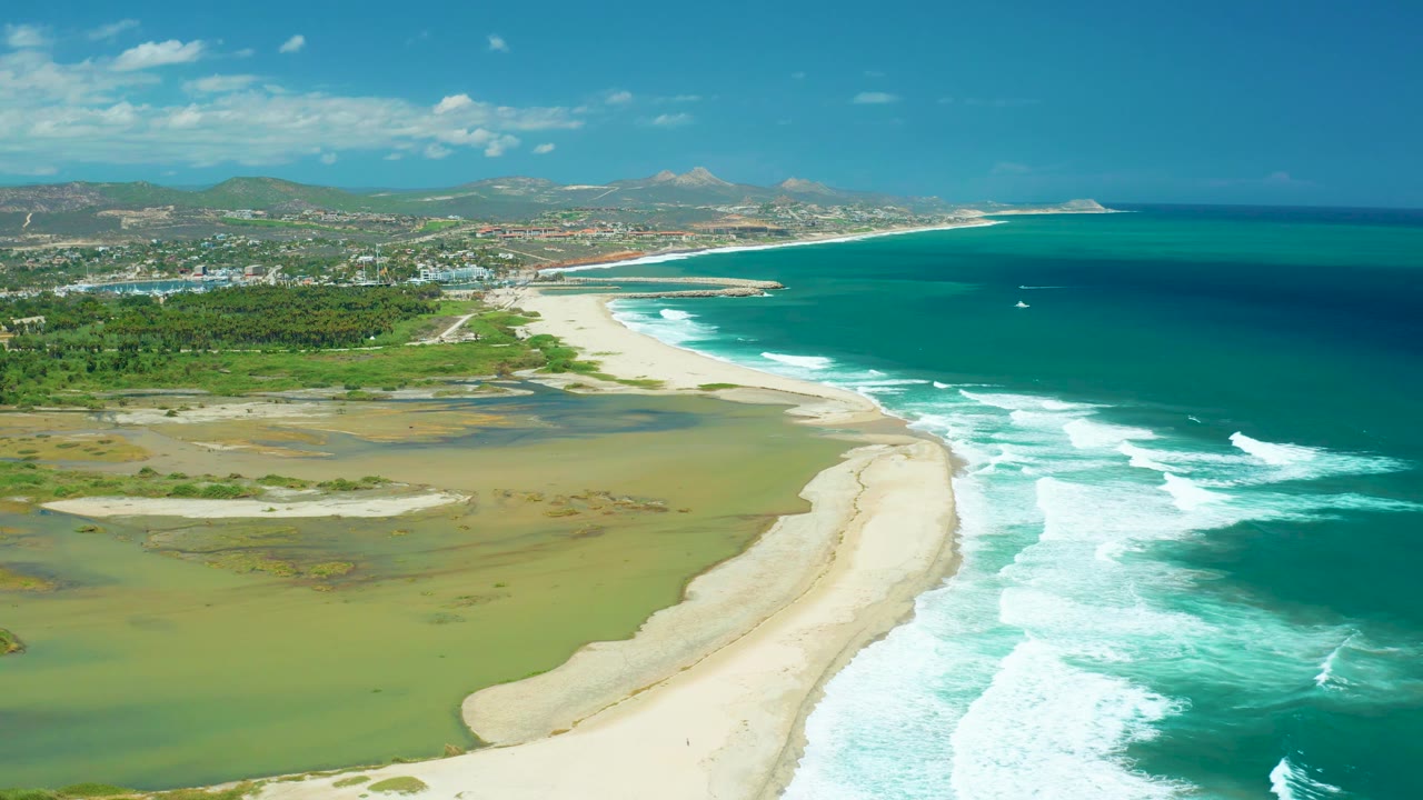 Wide aerial view in San Jose del Cabo with the sand spit centered between shallow lagoon water and rolling ocean waves, backed by low hills, scattered buildings, and no visible people.