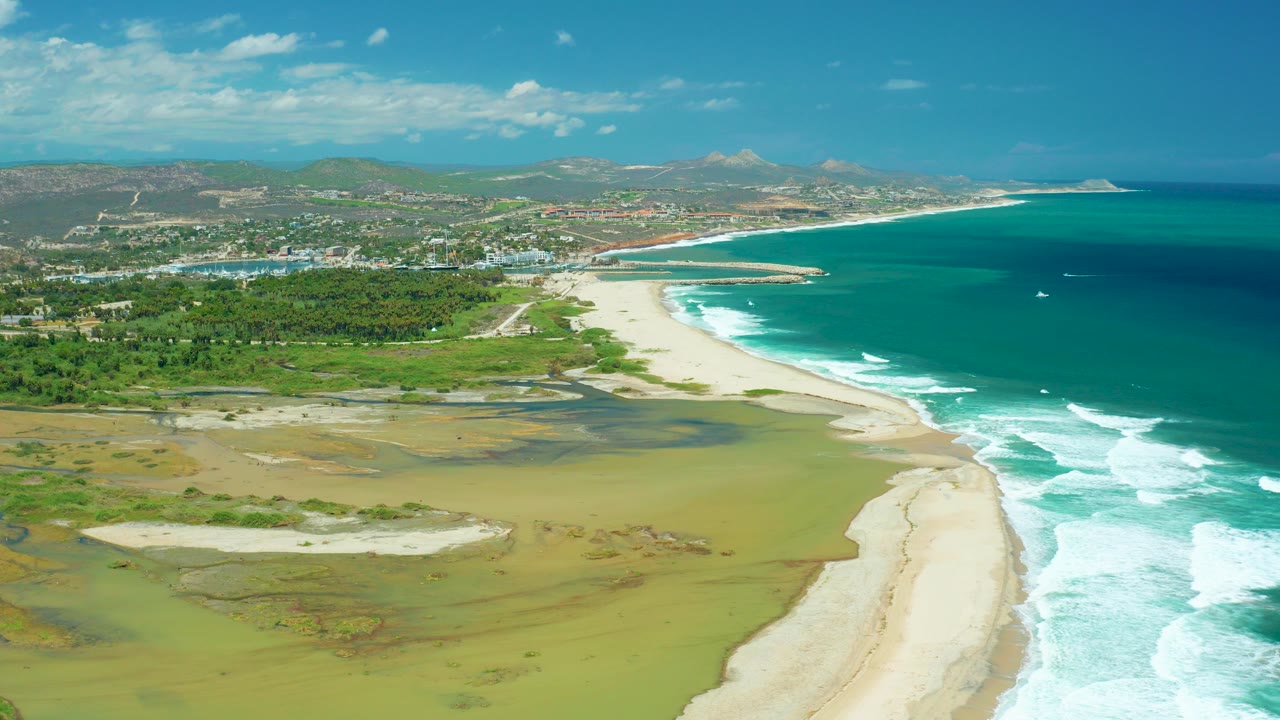 Aerial coastline with sandy beach, turquoise ocean, wetlands, town, hills, and blue sky.