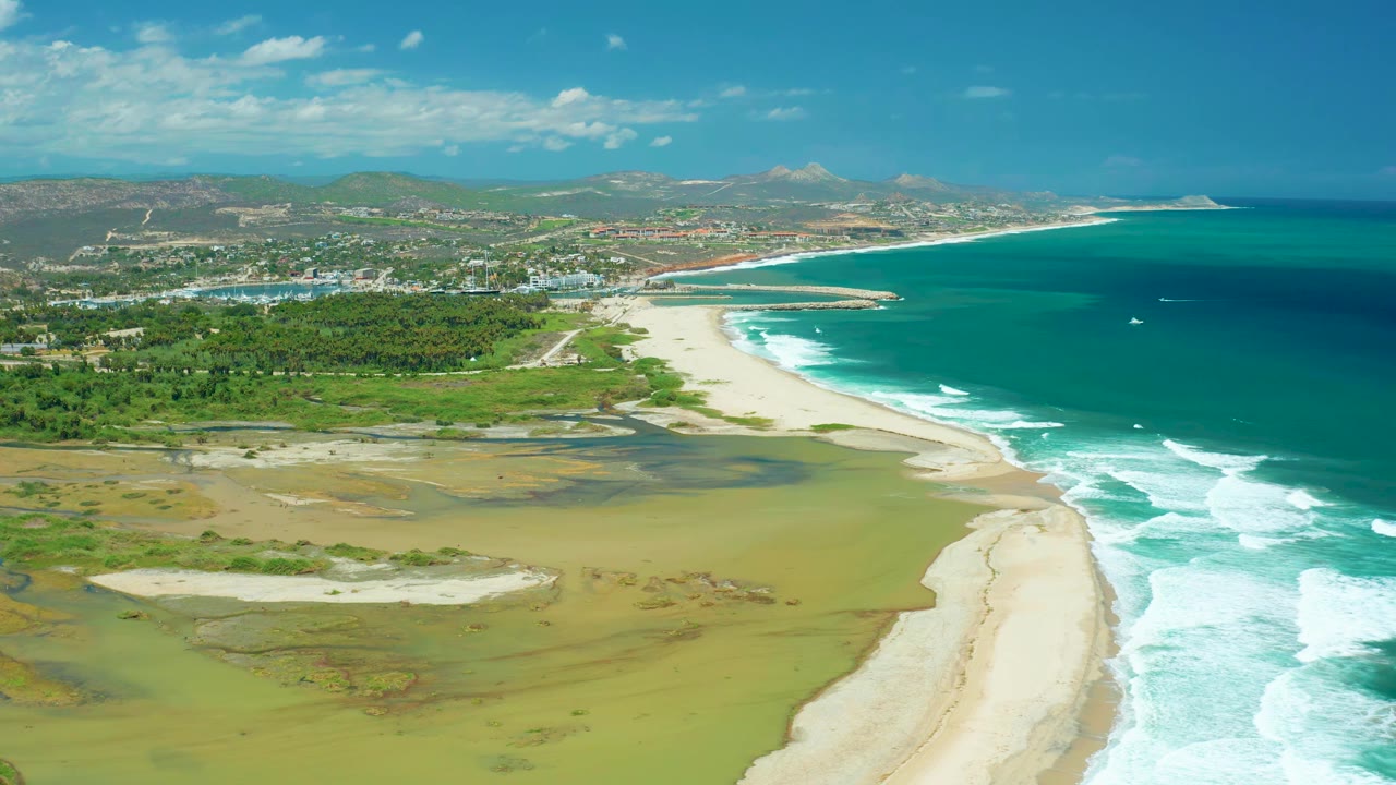 San Jose del Cabo coastline from above with the beach running diagonally through frame, ocean waves on the right, wetland pools on the left, distant buildings, hills, and no visible people.