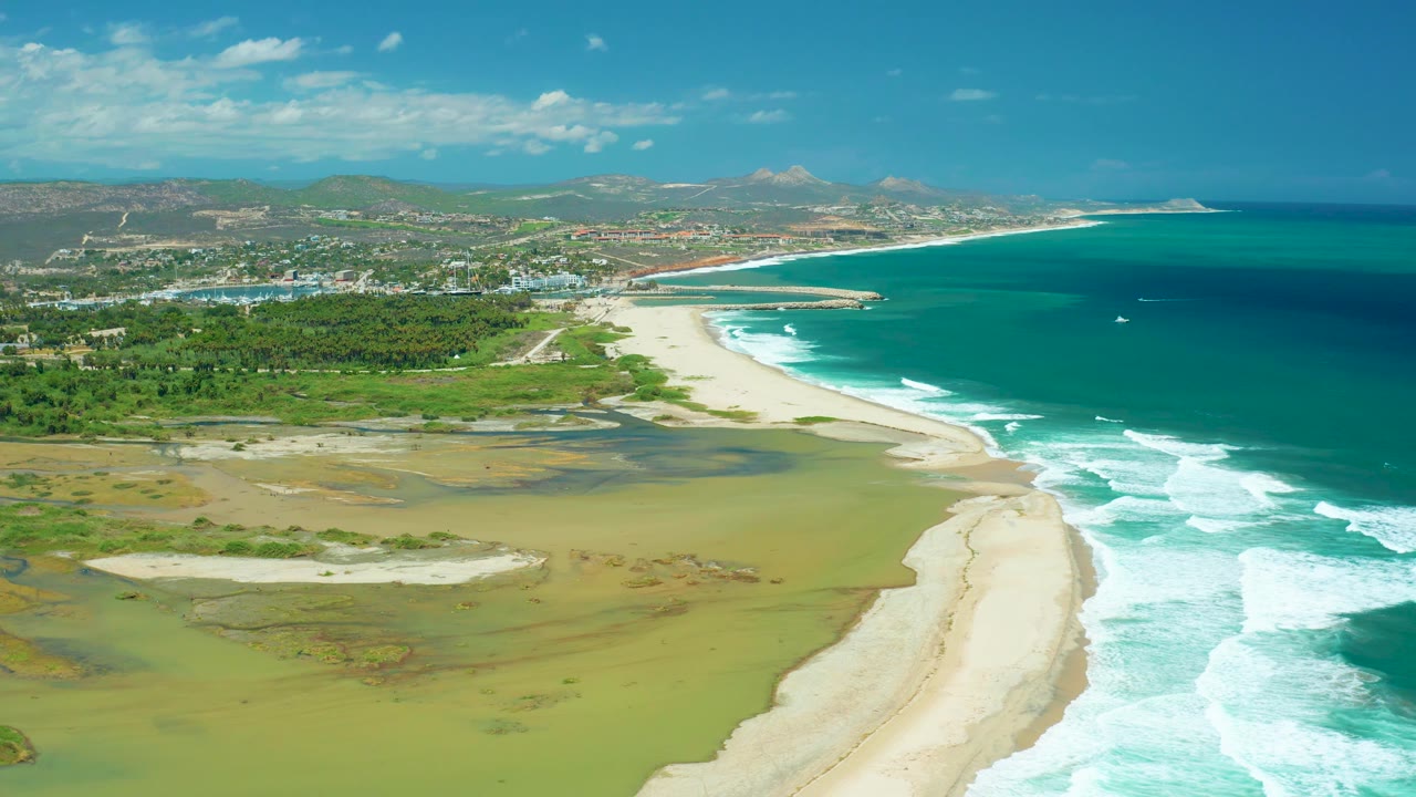 Wide San Jose del Cabo aerial view with broad wetland flats in the foreground, a pale beach separating inland water from ocean surf, distant development, hills, clouds, and no visible people.