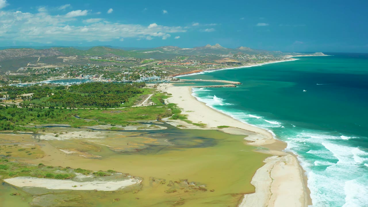 Wide aerial coastline with sandy lagoon, beach surf, turquoise water, greenery, and hills.