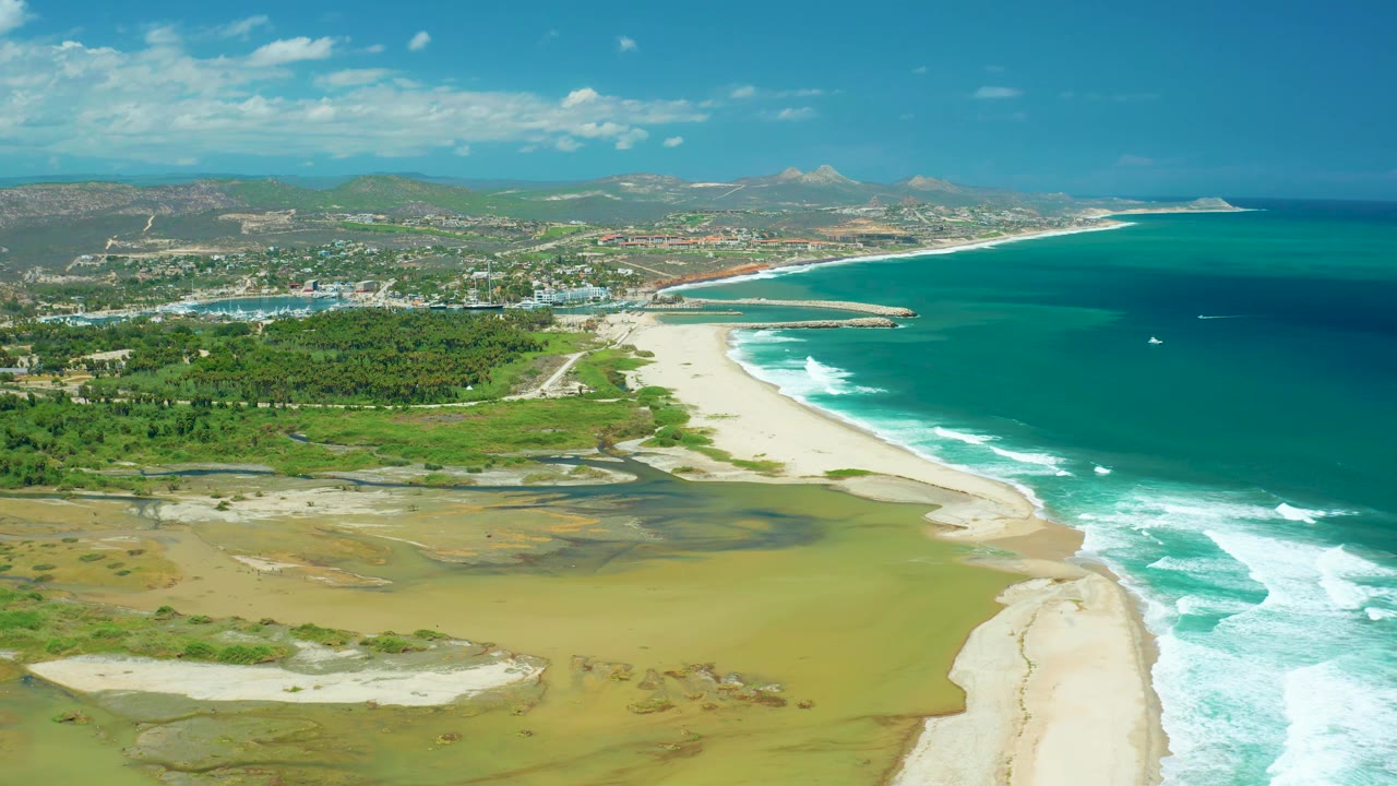 Aerial view in San Jose del Cabo where exposed lagoon sand fills the lower frame, green vegetation borders the beach, waves break along the right edge, and no visible people appear.