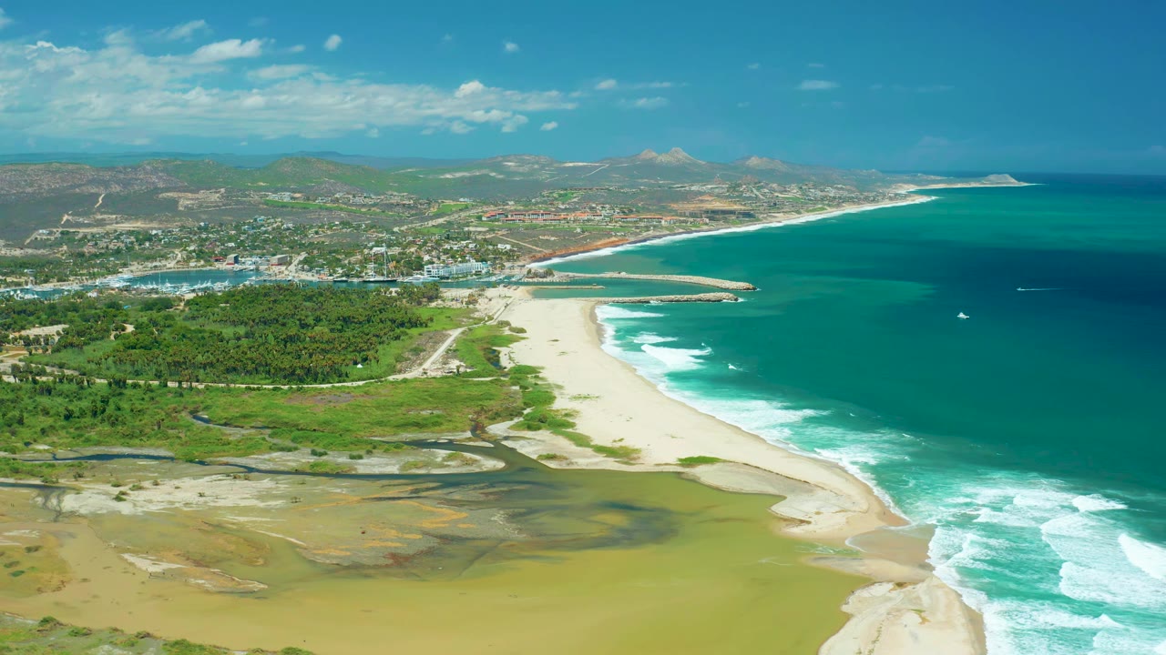 Aerial view in San Jose Del Cabo with exposed muddy lagoon shore in front, bright sand curving along the coast, white surf bands on turquoise water, inland greenery, distant buildings, hills, and no visible people.
