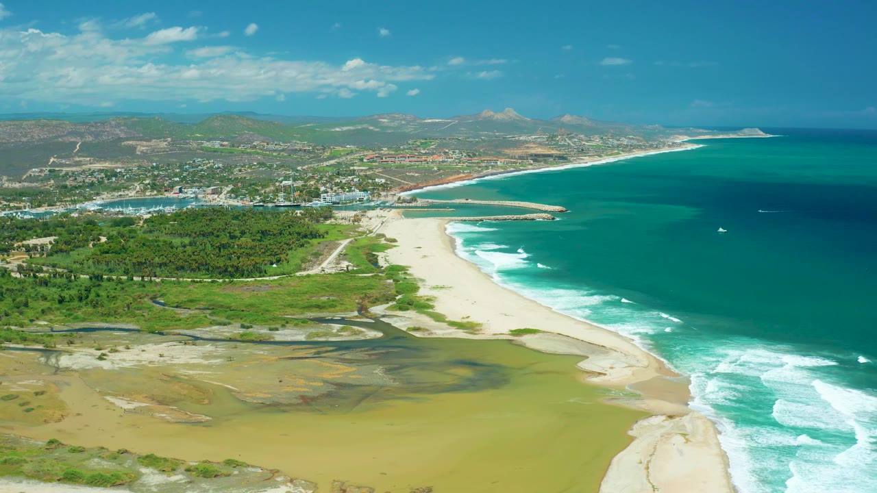 San Jose Del Cabo aerial shoreline frame with shallow yellow-green lagoon water and mudflat below, a wide sandy beach at center, ocean surf on the right, green vegetation inland, distant town, hills, and no visible people.