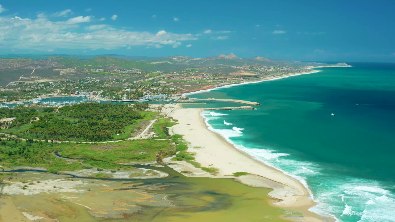 San Jose del Cabo coastline from above with surf rolling onto the sandy shore, green land in the foreground, a town beyond the beach, mountains on the horizon, and no visible people.