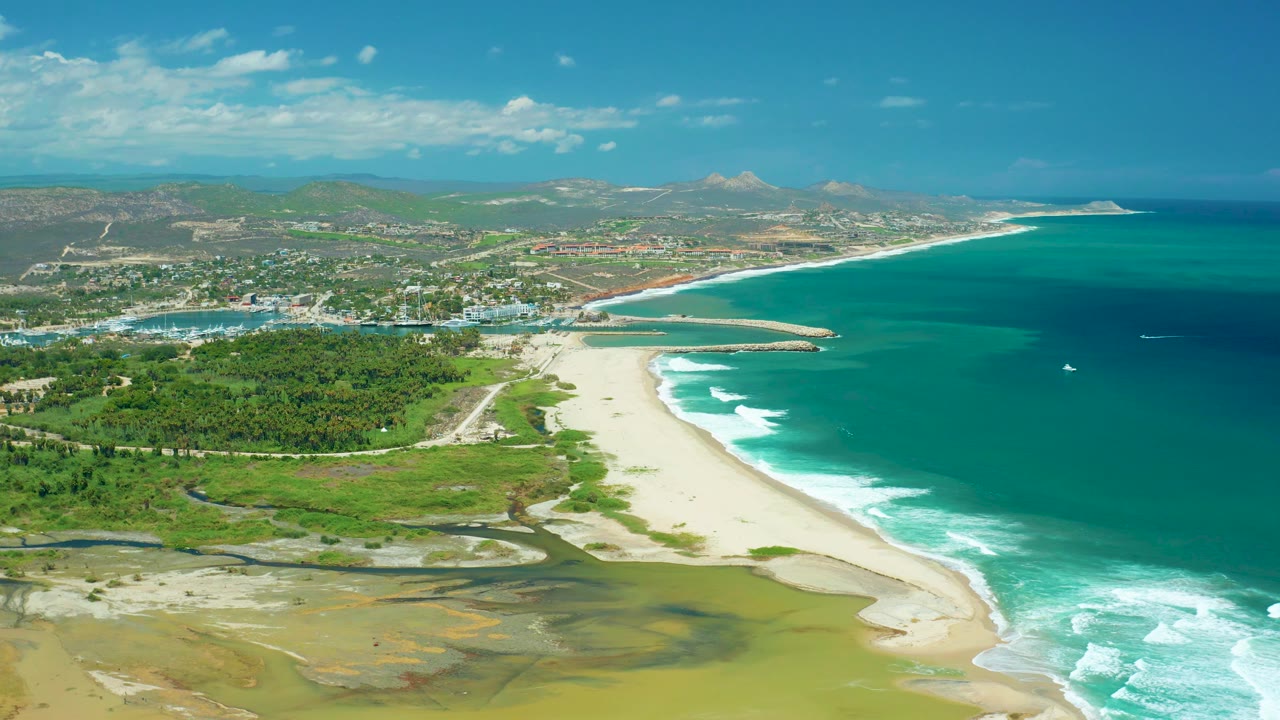 San Jose del Cabo aerial view with shallow water patterns in the foreground, a bright beach, white surf stripes, blue ocean, distant town and hills, and no visible people.