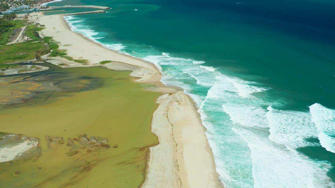 The San Jose del Cabo shoreline remains framed from above as white surf pushes along the beach beside the muted green estuary, with no visible people.
