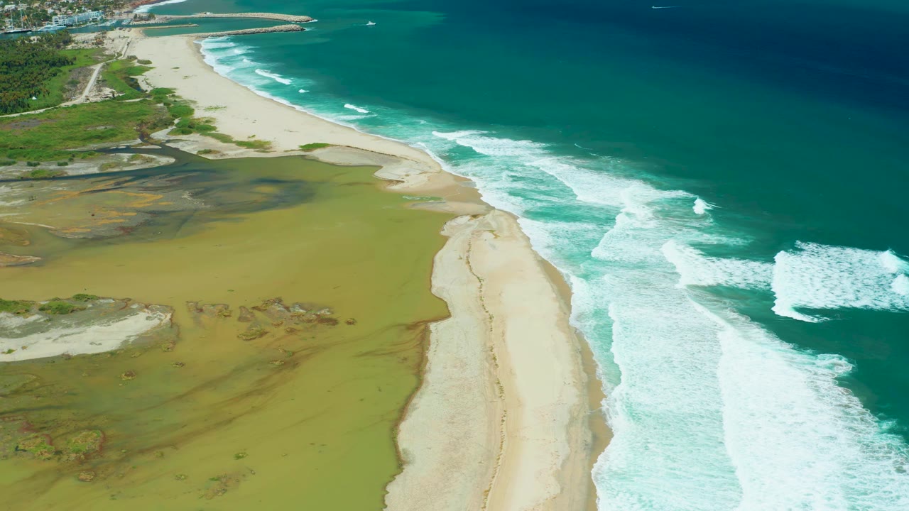 San Jose Del Cabo aerial frame showing a diagonal sandy beach, foamy waves advancing from the ocean, green-brown wetland flats inland, small distant buildings, and no visible people.
