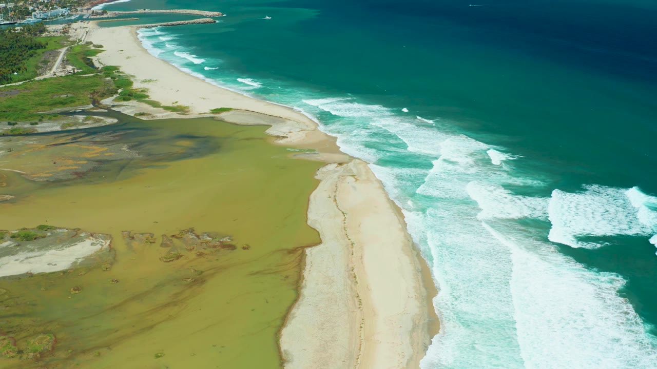 Aerial shoreline with surf washing along sand next to green-brown wetland water.