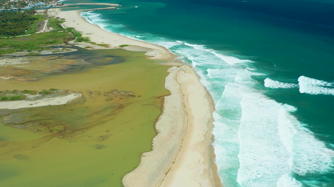 Aerial view in San Jose del Cabo showing green-brown wetland water beside a pale sand beach and turquoise ocean surf, with no visible people.