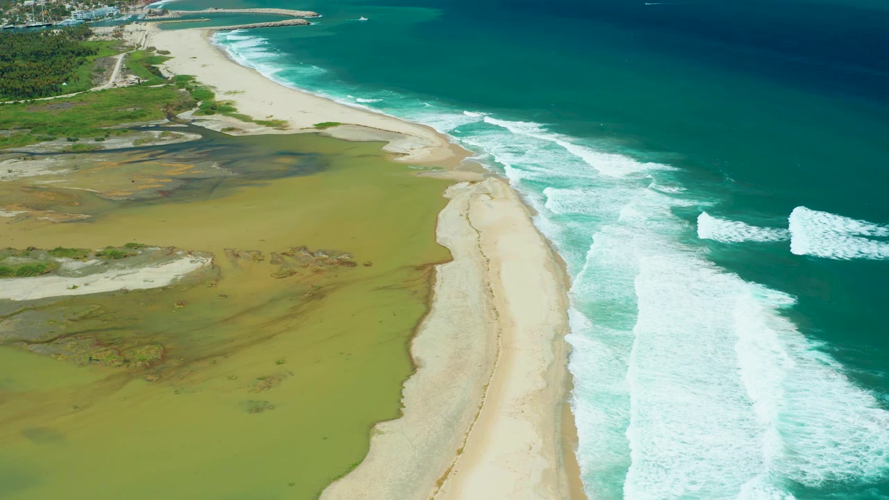 San Jose del Cabo aerial view with a narrow sandy strip dividing mottled wetland shallows from turquoise ocean and breaking white waves, with no visible people.