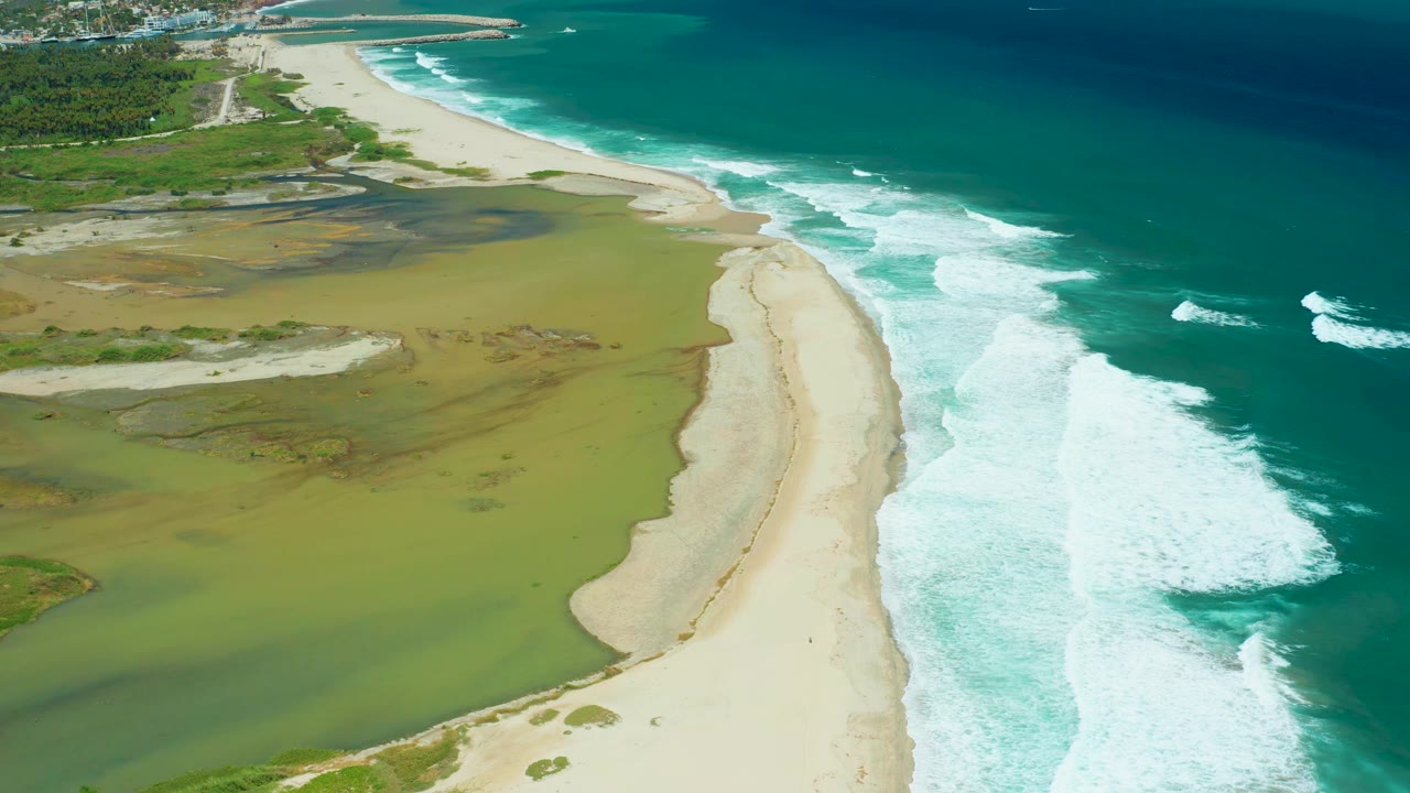 Aerial view in San Jose Del Cabo with the central sandbar curving past green lagoon water while white ocean foam advances along the right side, no visible people.