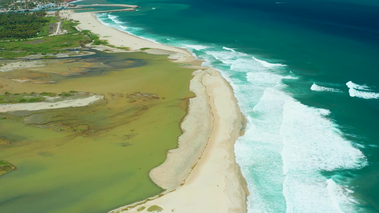 Aerial San Jose Del Cabo view with more lagoon surface in the left foreground, the pale beach running upward through frame, and ocean foam breaking to the right, no visible people.